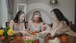 Beautiful young women happily eat pizza, putting aside their fresh vegetable salads, sitting at a table in the living