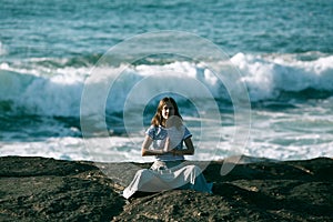 Beautiful young yoga woman meditates in the lotus position, sitting on the rocks on the Alantic coast. photo