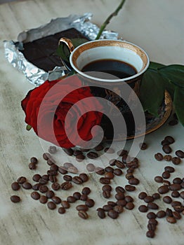 Vintage beautiful cup of hot coffee, bright red rose, green leaves, scattered coffee beans and dark chocolate bar on a wooden tabl