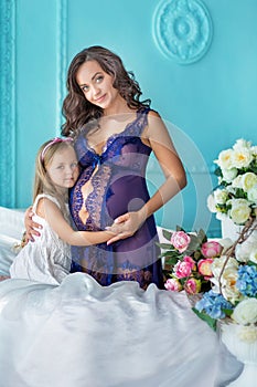 Beautifull young brunette pregnant woman in awesome purple dress close to blue sofa and cute flowers together with daughter.