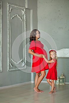 Beautifull young brunette pregnant woman in awesome purple dress close to blue sofa and cute flowers together with daughter.