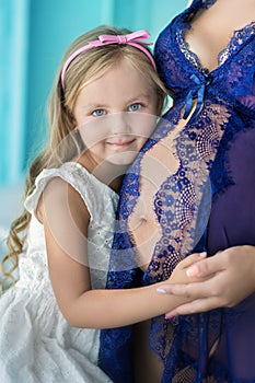 Beautifull young brunette pregnant woman in awesome purple dress close to blue sofa and cute flowers together with daughter.