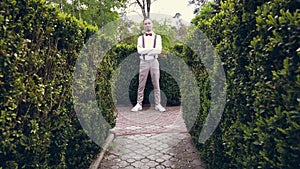 Among the beautifully cropped bushes in the park, a young man in a white shirt, trousers, stylish suspenders and bow tie