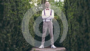 Among the beautifully cropped bushes in the park, a young man in a white shirt, trousers, stylish suspenders and bow tie