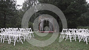 Beautifully decorated wedding reception on a green lawn near green fir trees with white chairs