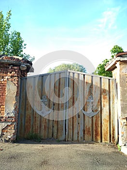 Beautifully light blue sky and greenery of young birches. View of the old rusty gray gate. At the gate are old anchor signs.