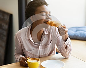 Beautuful young black woman enjoying her croissant and coffee with closed eyes at city cafe