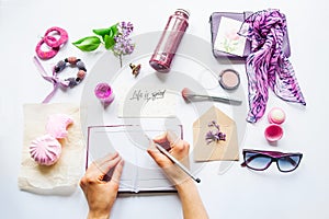 Beauty blog concept. Lilac colour. Female hands writing in notebook among styled accessories: sunglasses, bijouterie, cosmetics an