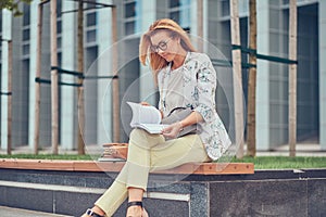 Charming blonde female in modern clothes, studying with a book, sitting on a bench in the park against a skyscraper.