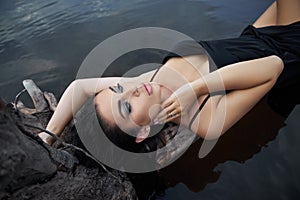 Beauty brunette woman in a black dress poses in a sea lake against a blue sky background. Long hair sexy woman and beautiful