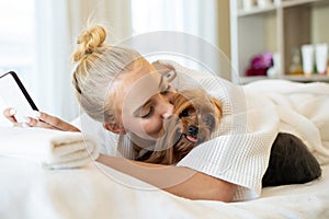 Beauty caucasian woman in pajamas using smart phone in the bed and kissing her cute small brown dog yorkshire terrier. Sleeping