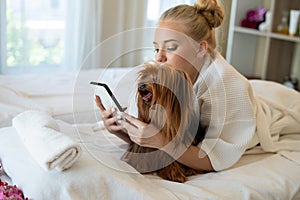 Beauty caucasian woman in pajamas using smart phone in the bed and kissing her cute small brown dog yorkshire terrier. Sleeping