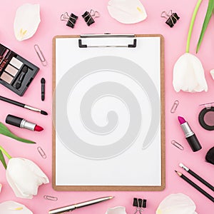 Beauty concept with clipboard, flowers, cosmetics and accessory on pink background. Top view. Flat lay. Feminine desk.