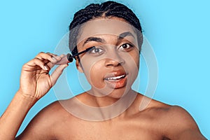 Beauty Concept. Young african woman isolated on blue applying mascara smiling concentrated close-up