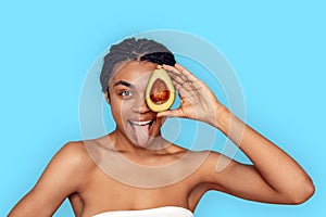 Beauty Concept. Young african woman isolated on blue covering eye with avocado showing tongue smiling playful