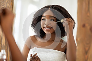 Beauty cosmetics for young skin. Sensual black woman applying regenerating serum onto her face near mirror indoors