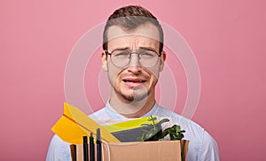 Beauty crying guy in a shirt and glasses holds a cardboard box with pens, plant and paper airplane