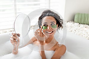 Beauty day and home skin care. Lovely young female applying patches under eyes, looking in mirror, lying in foamy bath