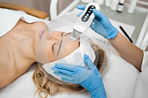 Beauty doctor with ultrasonic scraber doing procedure of ultrasonic cleaning of face. Cosmetology and facial skin care.