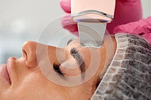 Beauty doctor with ultrasonic scraber doing procedure of ultrasonic cleaning of face. Cosmetology and facial skin care.
