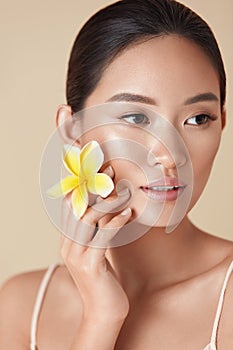Beauty Face. Flower And Model Close Up Portrait. Beautiful Asian Woman With Plumeria Looking Away Against Beige Background.