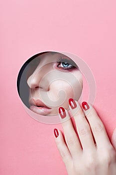 Beauty face red makeup plump lips red nails of a young girl in a round slit hole of pink paper. Woman with beautiful makeup plump