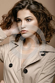 Beauty fashion model with clean skin and curly hair in biege cloak stretch with scarf on biege background