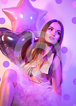 Beauty fashion model party Valentine`s Day girl with heart shaped air balloons, sitting on chair, laughing in neon light