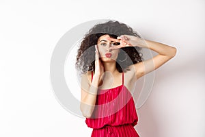 Beauty and fashion. Stylish curly-haired woman in red dress showing v-sign on eye and pucker lips, kissing you, standing