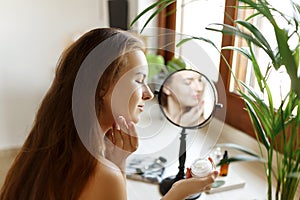 Beauty girl looking at mirror while touching her face and applying cream, wrinkles during morning beauty routine. Happy