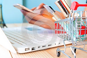 Beauty items and cosmetics in shopping cart with woman shopping online on smartphone