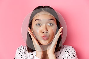 Beauty and lifestyle concept. Close-up of adorable asian woman touching face, pucker lips in kiss, standing over pink