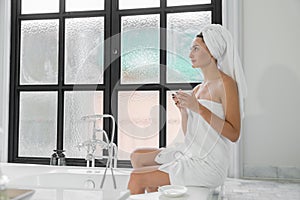 A beauty-loving woman wearing a towel with a healthy, smooth body looking at a round mirror in a white bathtub in a bathroom