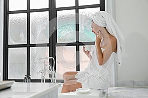 A beauty-loving woman wearing a towel with a healthy, smooth body looking at a round mirror in a white bathtub in a bathroom
