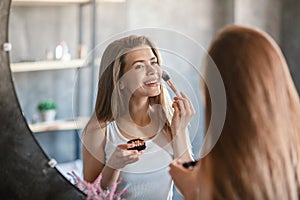 Beauty and makeup. Pretty young woman applying blusher on her face near mirror at home