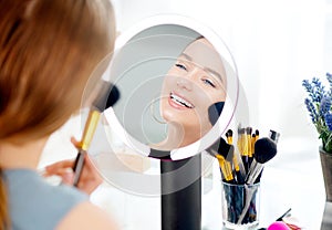 Beauty model girl applying makeup. Beautiful young woman looking in the mirror and applying cosmetic with a big brush