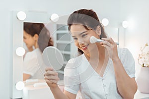 Beauty model girl applying makeup and smiling. Beautiful young woman looking in the mirror and applying cosmetic with a big brush