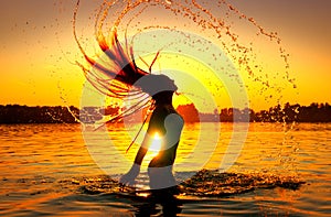 Beauty model girl splashing water with her hair. Girl silhouette over sunset sky. Swimming and splashing on summer beach