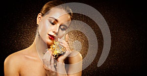 Beauty Model with Golden Makeup holding Gold Jewelry. Face Skin Care and Luxury Cosmetics. Shining Glitter Dark Background