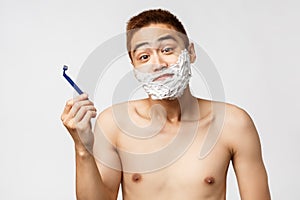 Beauty, people and hygiene concept. Clueless handsome asian teenage male trying shave beard first time, apply shaving