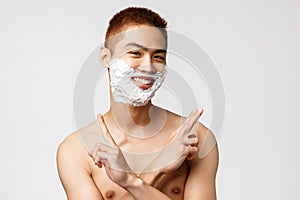 Beauty, people and hygiene concept. Portrait of funny teenage asian guy shaving first time, apply cream on beard and