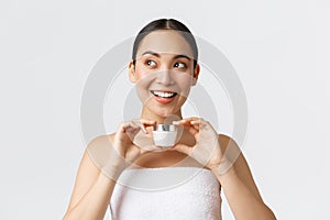 Beauty, personal care, spa salon and skincare concept. Excited beautiful asian woman in bath towel showing face cream