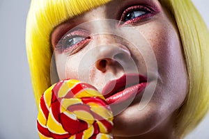 Beauty portrait of calm cute young female model with freckles, red makeup and yellow wig, holding colorful candy stick near face