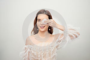 Beauty portrait of a cheerful young topless woman removing face make-up with a cotton pad isolated over gray background