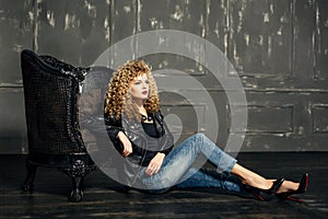 Beauty portrait of curled hair blonde girl sit on the floor leaning on the chair. studio with dark background