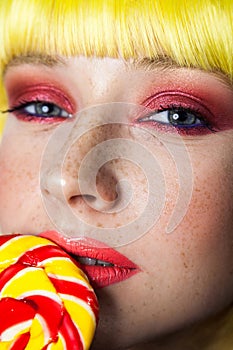 Beauty portrait of cute young girl with freckles, red makeup and yellow wig, holding colorful candy stick on lips and looking at