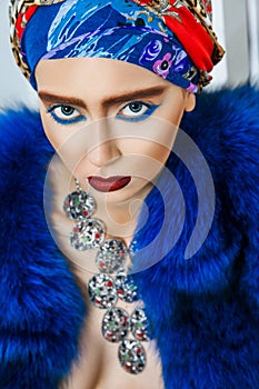 Beauty portrait of fashion model with colored headwear, blue fur coat red eyebrow and lips makeup and necklace.