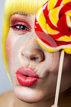 Beauty portrait of funny cute young female model with freckles, red makeup and yellow wig, holding colorful candy stick, kissing