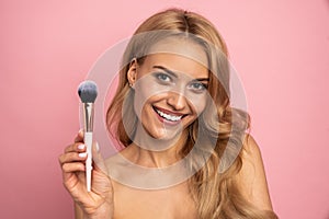 Beauty portrait of a lovely young woman with long blonde hair standing isolated over pink background, holding makeup brush