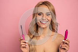 Beauty portrait of a lovely young woman with long blonde hair standing isolated over pink background, showing eye mascara